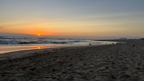 Una playa con el sol poniéndose sobre el océano, terreno arenoso y algunas personas caminando.