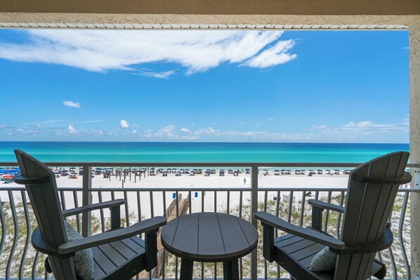 A balcony with two Adirondack chairs and a small table, overlooking a beach with umbrellas and people. - Crystal Sands 309a - Gulf Front, Free Beach Service, Heated Pool, Sleeps 9 (Destin)