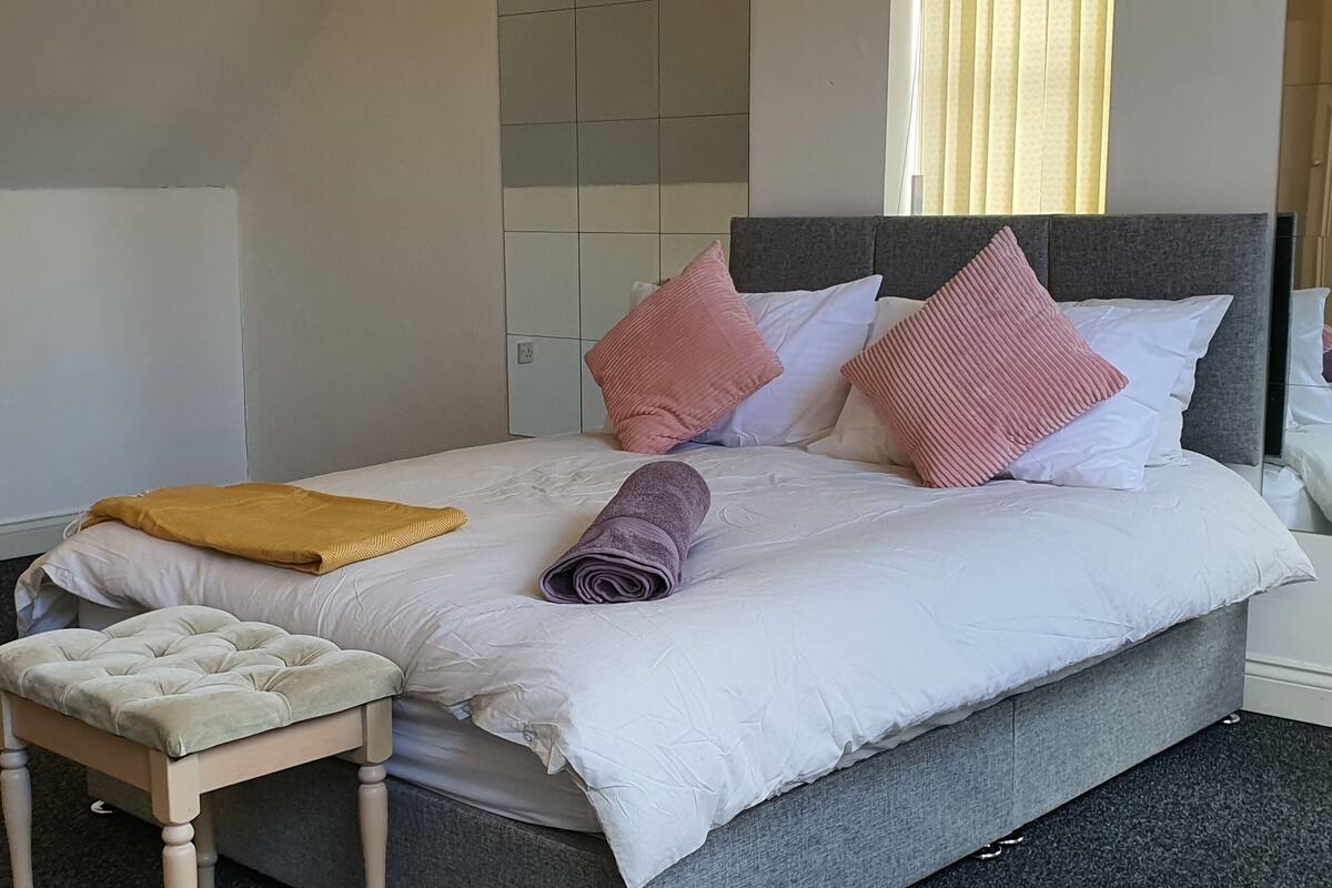 Ein ordentlich bezogenes Bett mit grauem Kopfteil, einer Holzbank und einer gelben Tagesdecke.