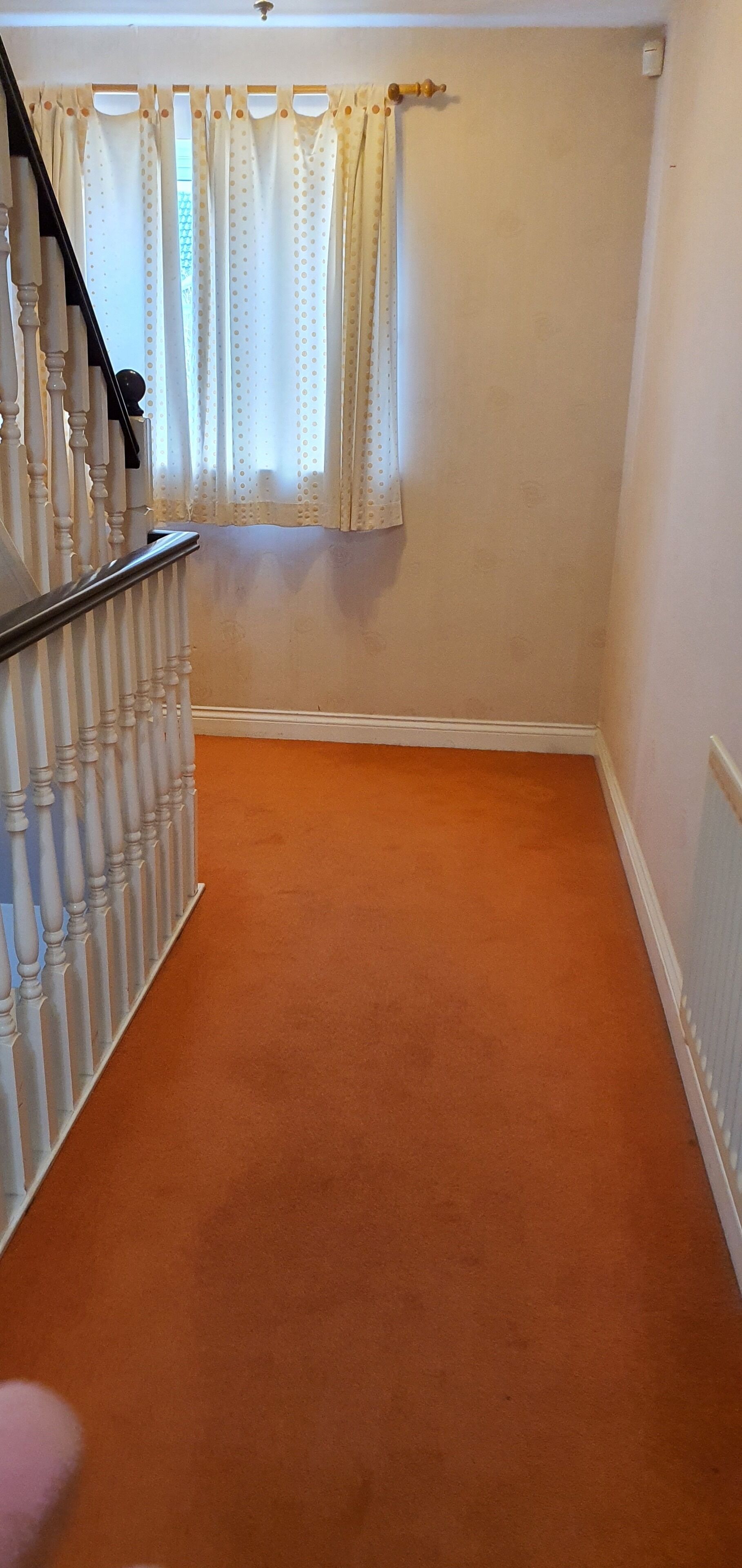 A narrow hallway with a carpeted floor, a staircase with white balusters, and a window with polka dot curtains.