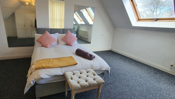 Une chambre avec un grand lit, un banc, un miroir et une lucarne.