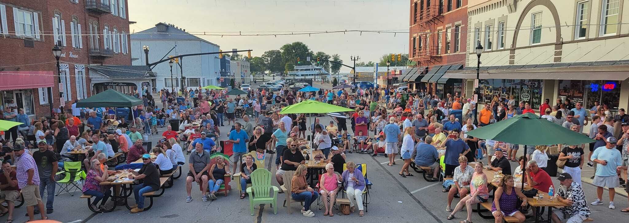 Une rue animée où l’on voit des gens assis à des tables et des chaises, probablement lors d’un événement en plein air ou dans un marché.