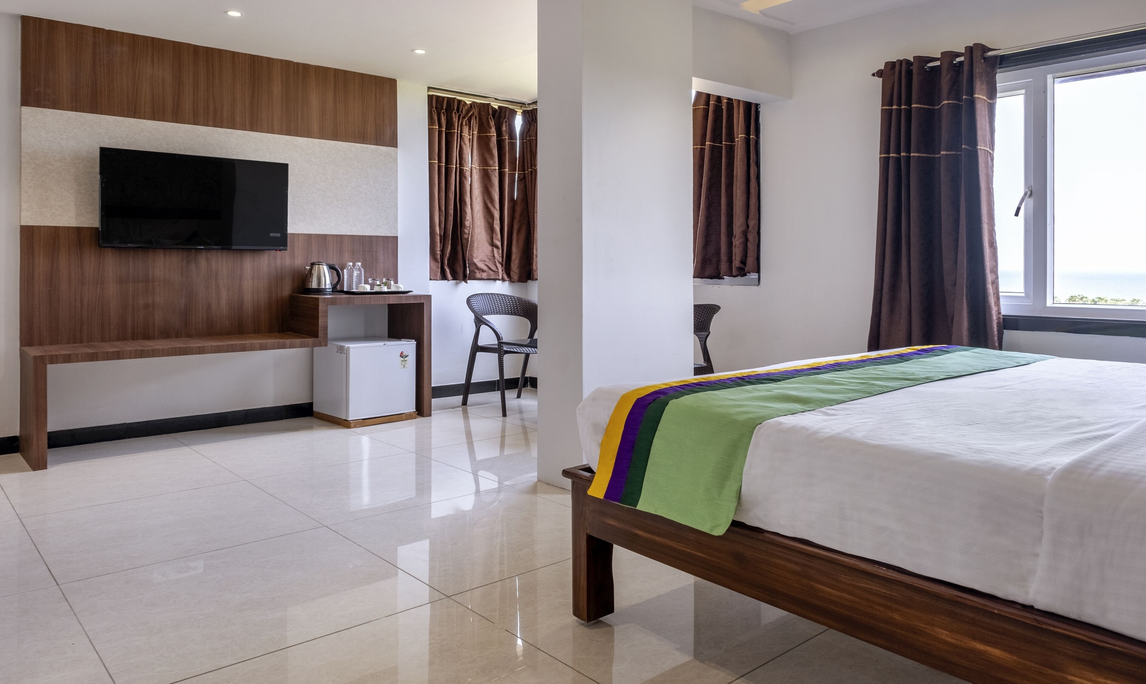 A hotel room with a bed, a TV mounted on the wall, a small fridge, and a desk with a kettle and bottles.
