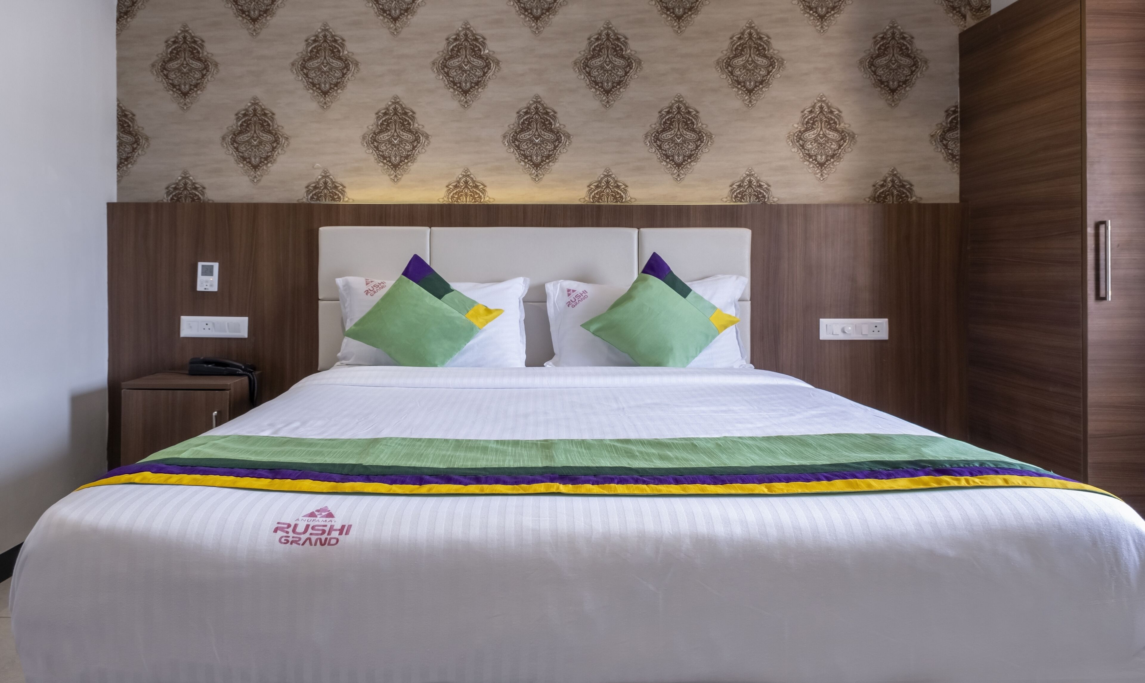 A hotel room with a large bed, two nightstands, a wooden wardrobe, and a patterned ceiling.