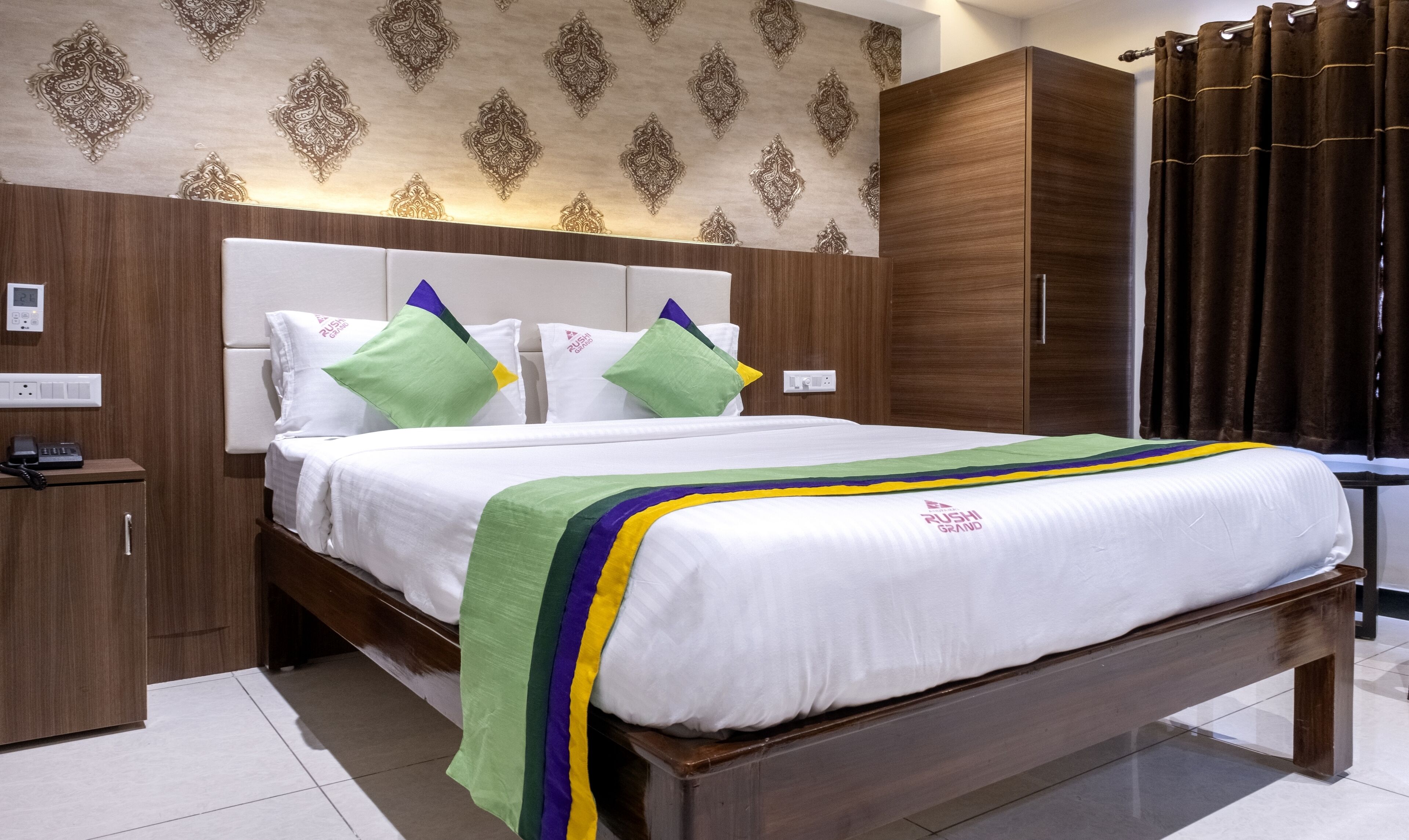 A hotel room with a large bed, a wooden wardrobe, a bedside table with a phone, and a wall with patterned wallpaper.