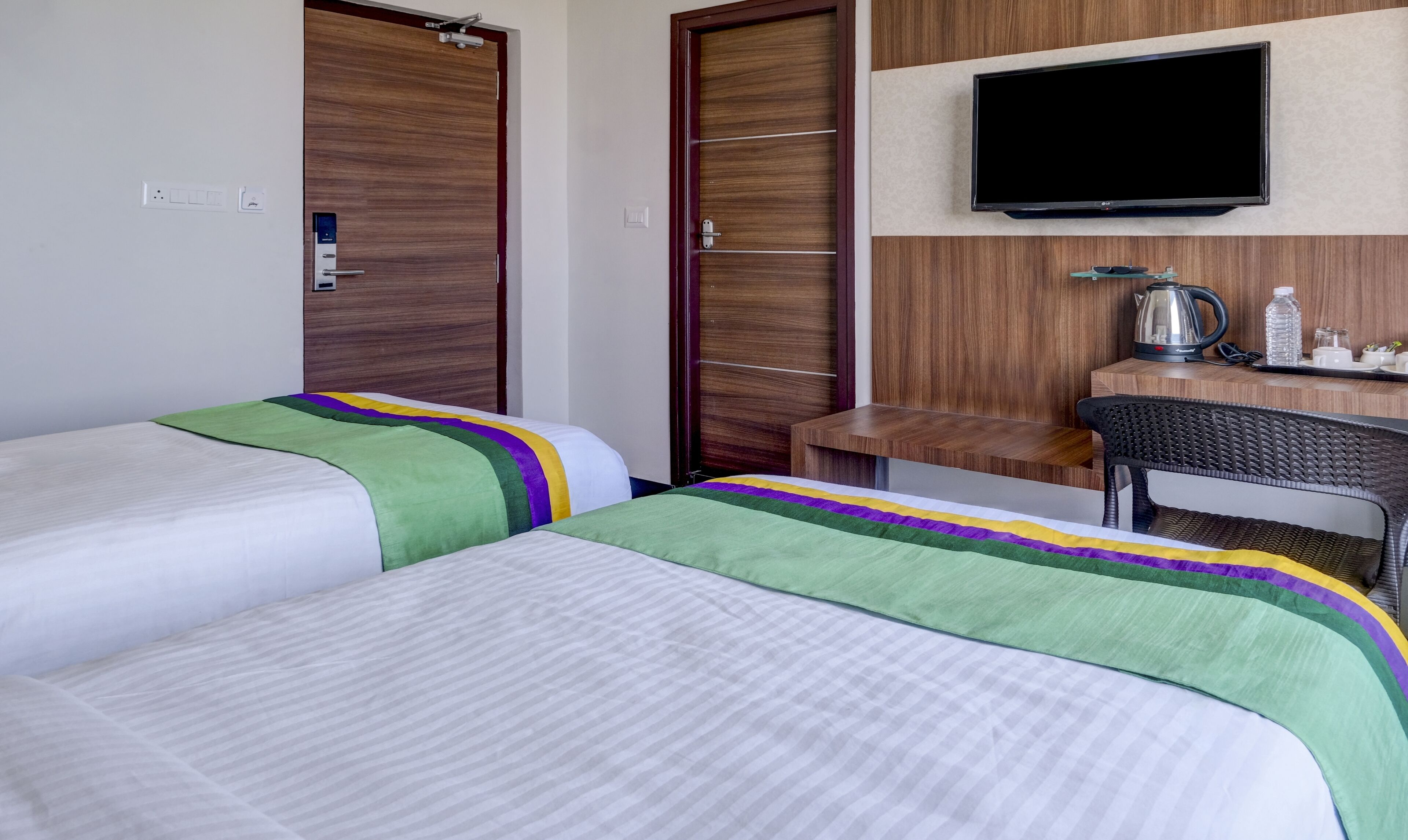 A hotel room with two beds, a TV, a desk with a kettle, and a chair.