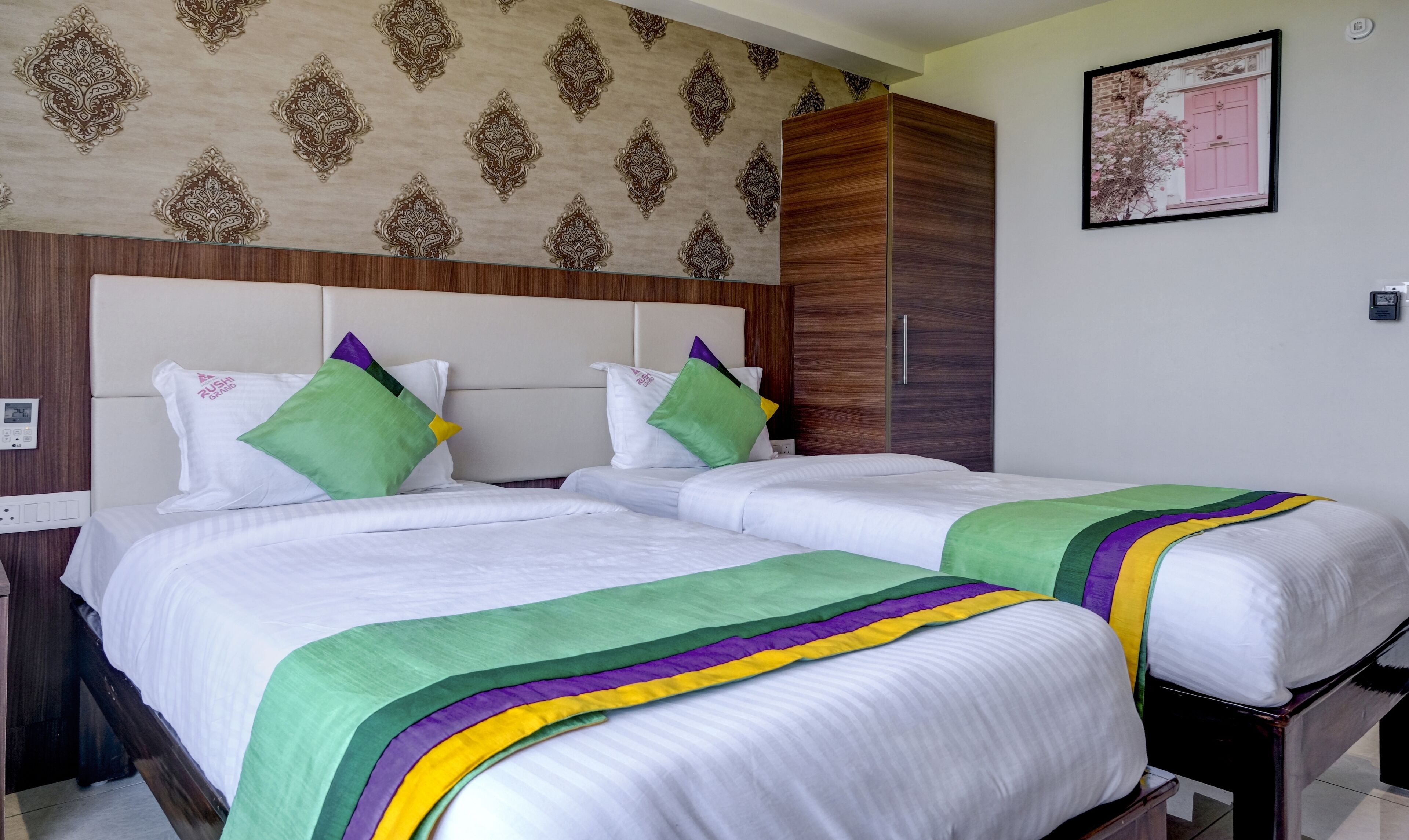 A hotel room with two beds, a wooden wardrobe, a patterned wallpaper, and a framed picture on the wall.