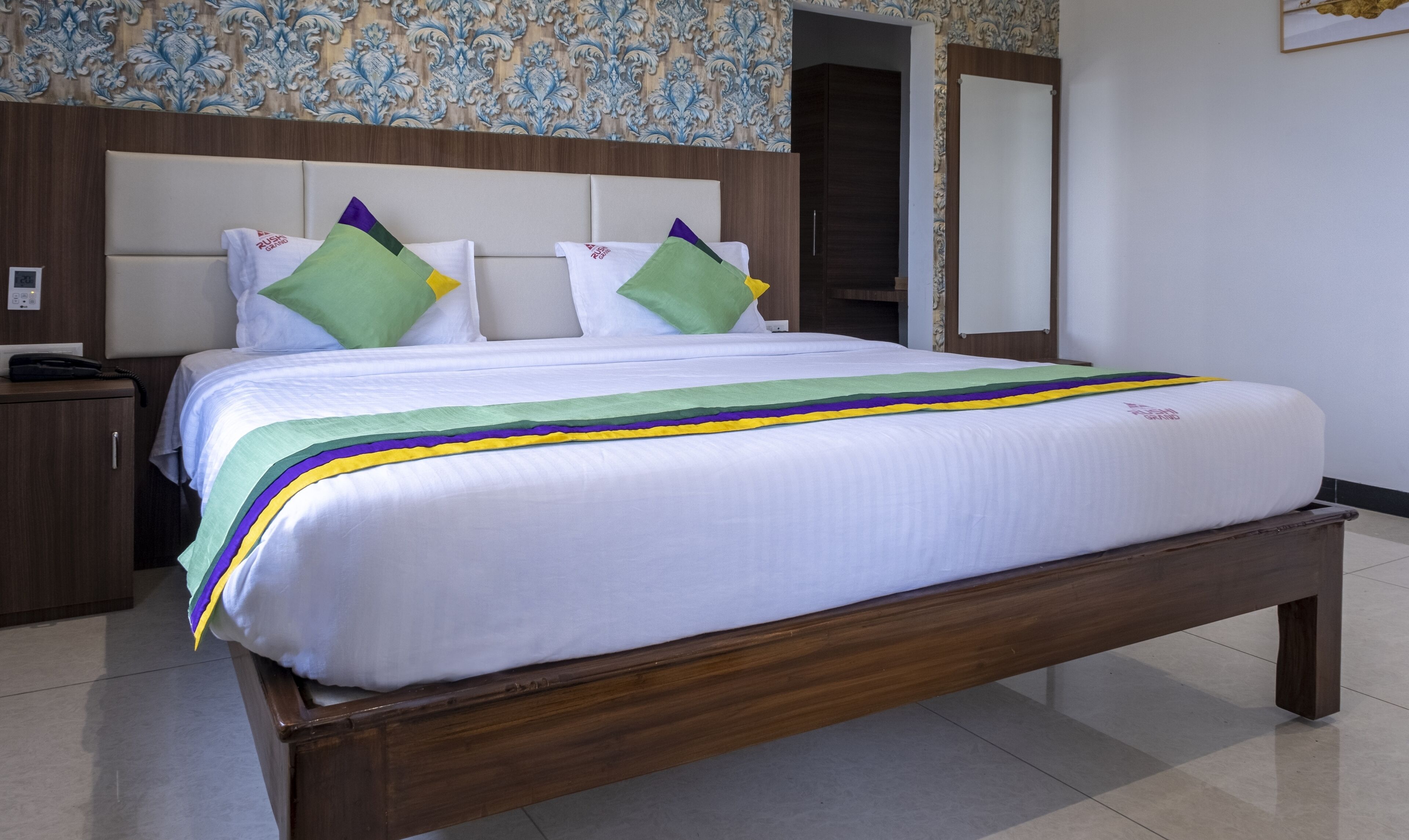 A hotel room with a large bed, a wooden headboard, a bedside table, and a wall with a decorative pattern.