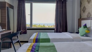 Habitación de hotel con dos camas, un escritorio y una silla. Cuenta con una ventana con cortinas y vistas al mar y a los árboles.