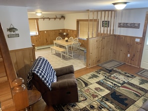 A cozy living room with a brown sofa, a patterned rug, wooden walls, a dining area with a table and chairs, and a 'Welcome' sign on the wall.