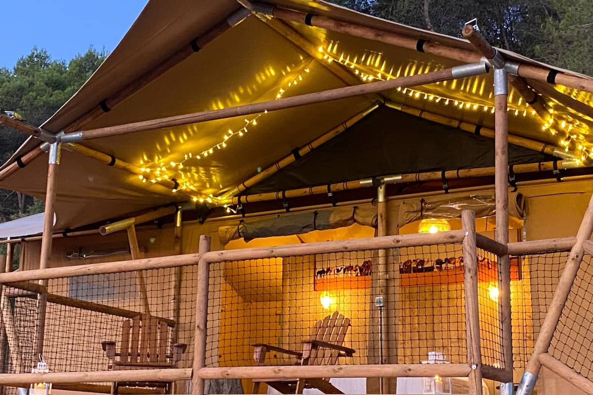 Una cabaña de madera con porche cubierto, luces colgantes y un patio cercado con una silla y una mesa.
