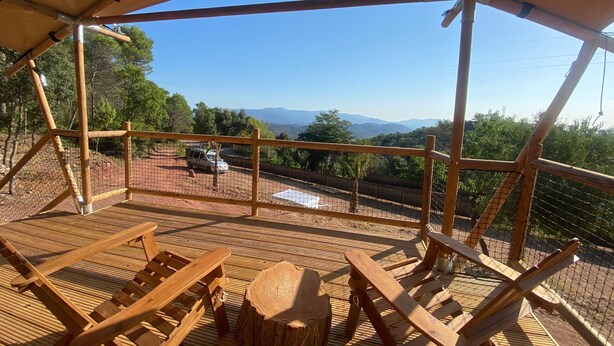 A wooden deck with lounge chairs and a table, overlooking a mountainous landscape. - Glamping VALL de CODÓ (Terrades)