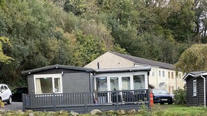 Un chalet au bord de la rivière, doté d’une terrasse et entouré d’une végétation luxuriante, avec une rivière qui serpente à proximité.