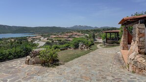 Un camino de piedra conduce a un edificio de piedra con techo rojo, con vistas a un pueblo costero con casas y una masa de agua.