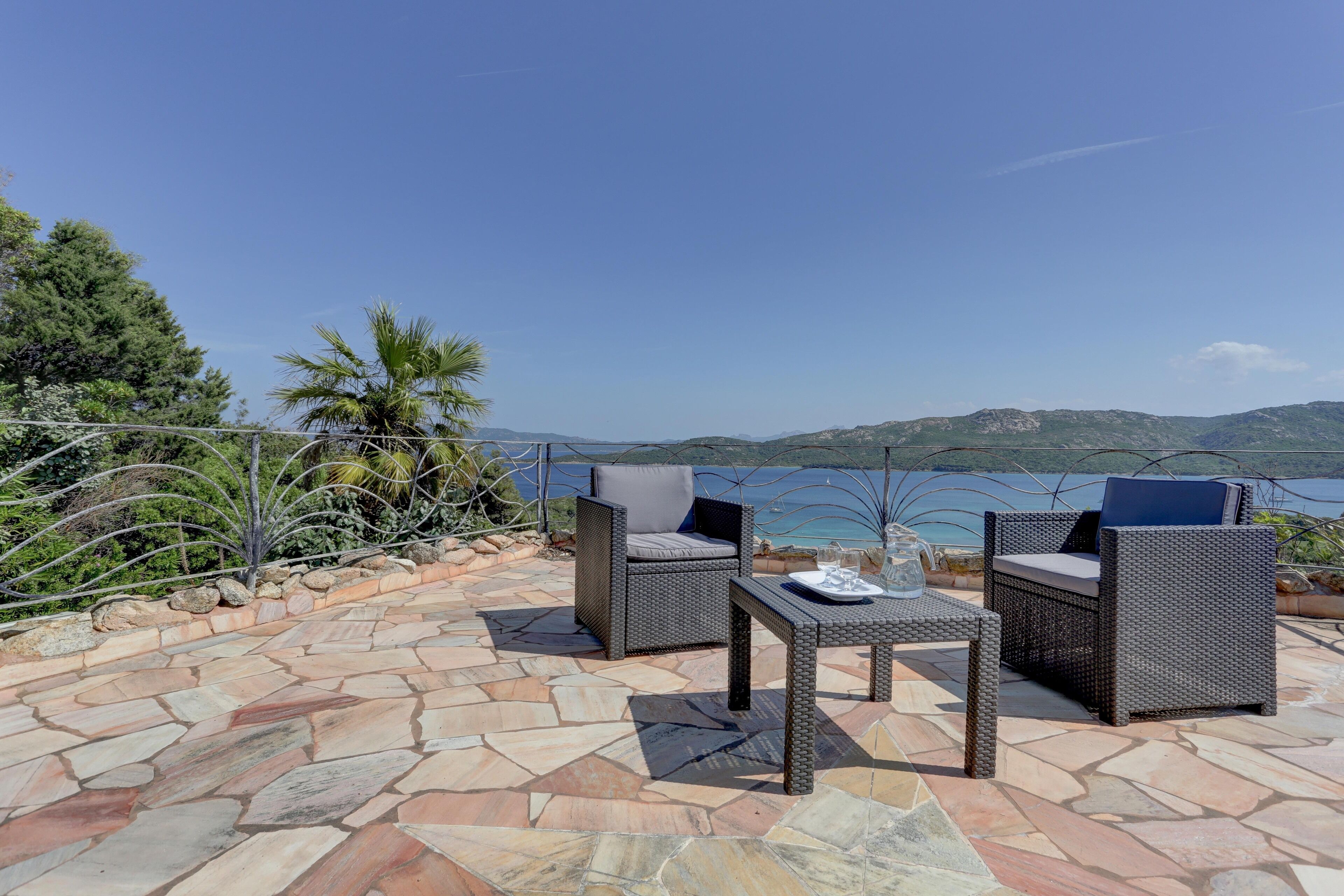 An outdoor patio with stone flooring, two wicker chairs, a small table, and a view of the sea and hills.