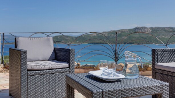Un espacio exterior con asientos de mimbre y una mesa dispuesta para dos personas, con vistas panorámicas al mar y las colinas.