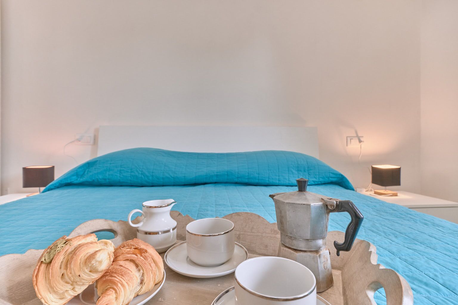 A bed with a blue bedspread and pillows, a tray with croissants, cups, and a coffee pot on a wooden table.
