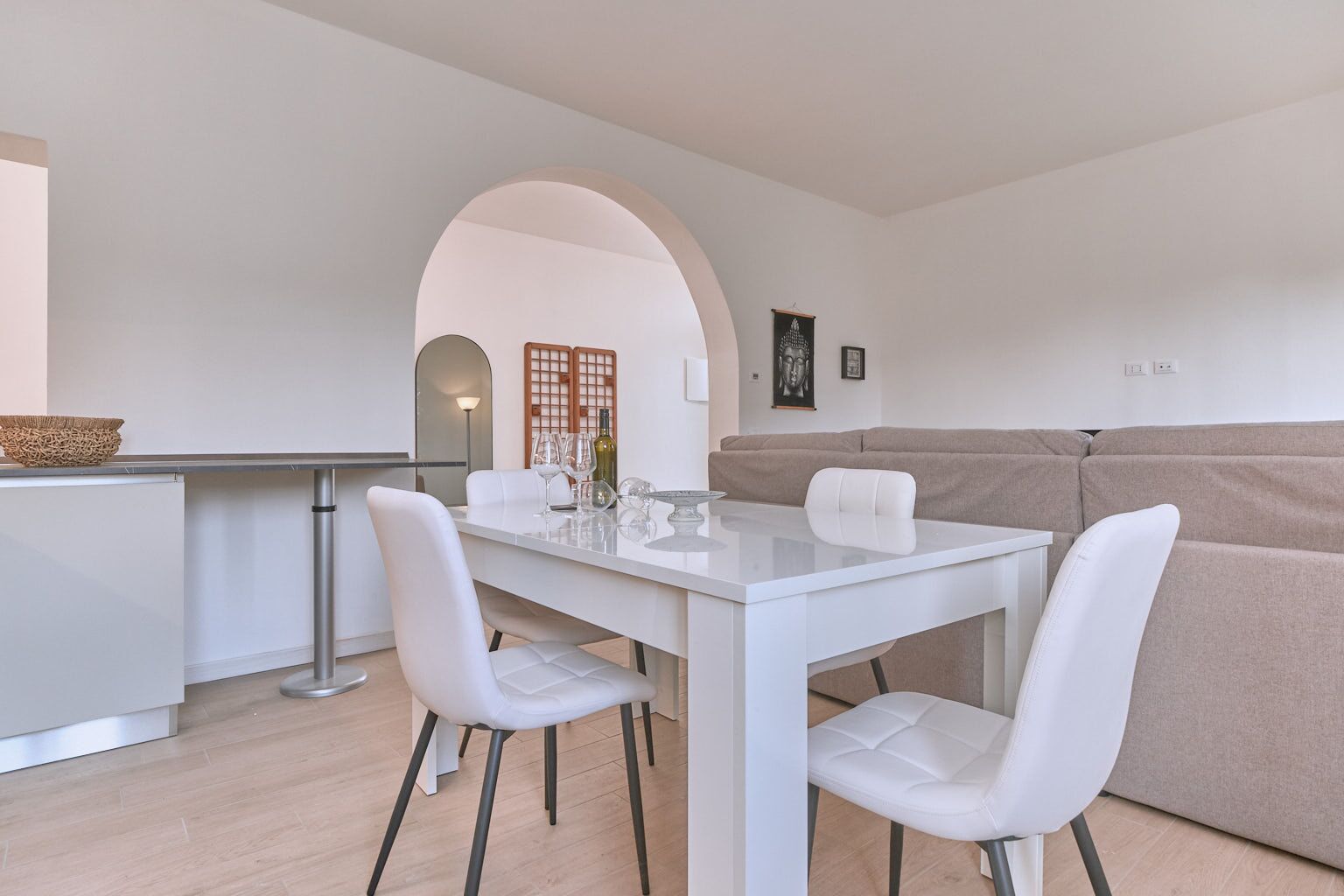 Een moderne eetruimte met een witte tafel en stoelen, een beige bank, en een keuken op de achtergrond.