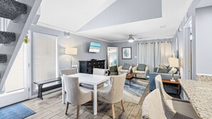 A modern living room with a dining area, a TV, and a large window. - Sea Cabins 9B - Heated Community Pool, Private Beach Access, Bikes Included! (Miramar Beach)