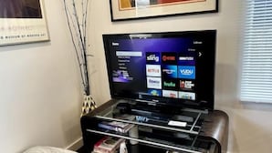 A flat-screen TV on a stand with a shelf below displaying books and DVDs. - Modern Ranch near Knapp Center, Stadium, Downtown (Des Moines)