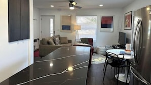 A modern living room with a dark marble floor, a sofa, a dining table with chairs, a ceiling fan, and a refrigerator. - Modern Ranch near Knapp Center, Stadium, Downtown (Des Moines)