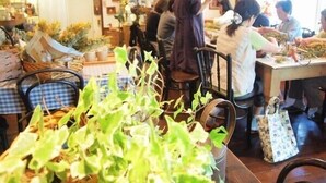 A restaurant with tables set for dining, a person standing by the entrance, and various plants and items on shelves.