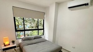 A bedroom with a bed, a nightstand with a lamp, a window with a view of greenery, and an air conditioning unit on the wall. - Modern 302 apartment w/AC +WiFi + Parking (Cali)