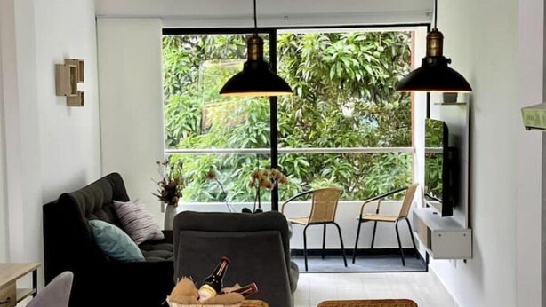 A modern living room with a sofa, dining table, and chairs. There is a large window with a view of greenery outside. - Modern 302 apartment w/AC +WiFi + Parking (Cali)