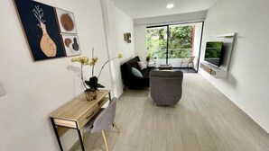 A modern living room with a sofa, a coffee table, and a TV mounted on the wall. There is a wooden desk with a plant on it, and a window with a view of greenery outside. - Modern 302 apartment w/AC +WiFi + Parking (Cali)