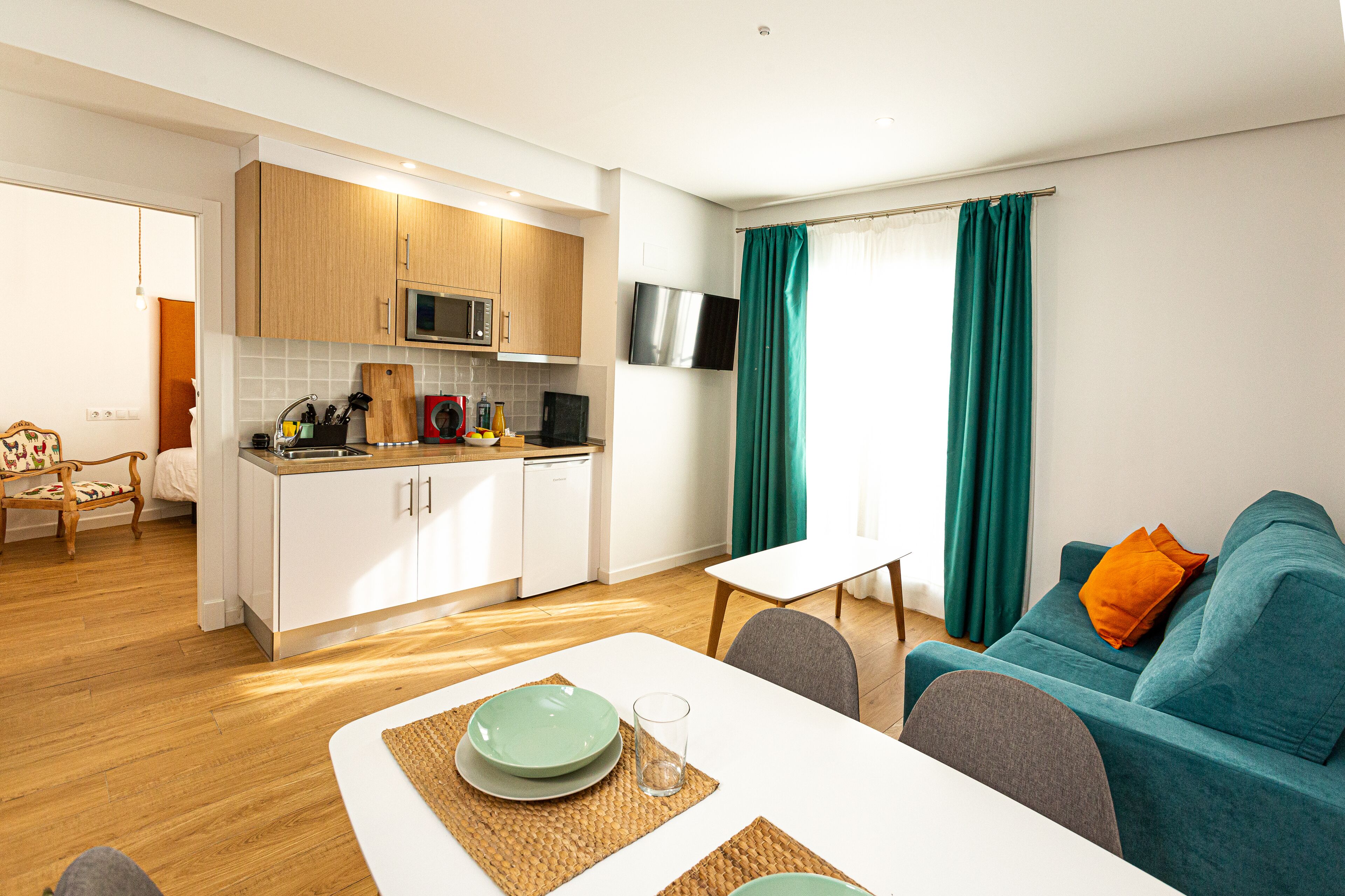 A modern kitchen with white cabinets, a microwave, and a dishwasher. There is a dining area with a white table and chairs, and a teal sofa with an orange pillow.