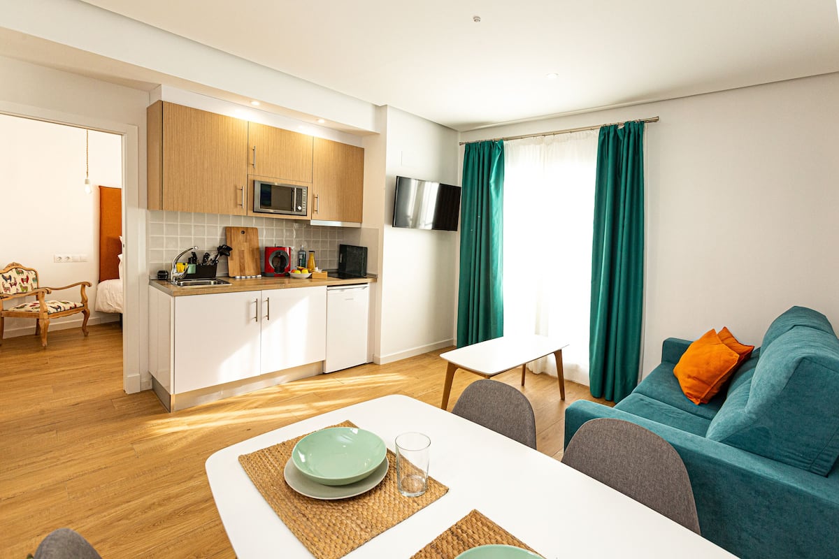 A modern kitchen with white cabinets, a microwave, and a dishwasher. There is a dining area with a white table and chairs, and a teal sofa with an orange pillow.