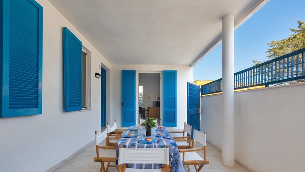 Un espacio de comedor al aire libre cubierto con una mesa dispuesta para cuatro, rodeado de contraventanas azules, paredes blancas y un suelo de baldosas.