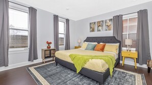 A bedroom with a large bed, two bedside tables, a nightstand, a window with curtains, and artwork on the wall.
