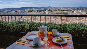 Une table dressée avec une théière, des tasses et une assiette de nourriture, placée sur un balcon donnant sur un paysage urbain.