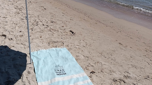 Une plage avec un parasol bleu sur lequel on peut lire « SARD GUEST HOUSE » et une serviette assortie posée sur le sable.