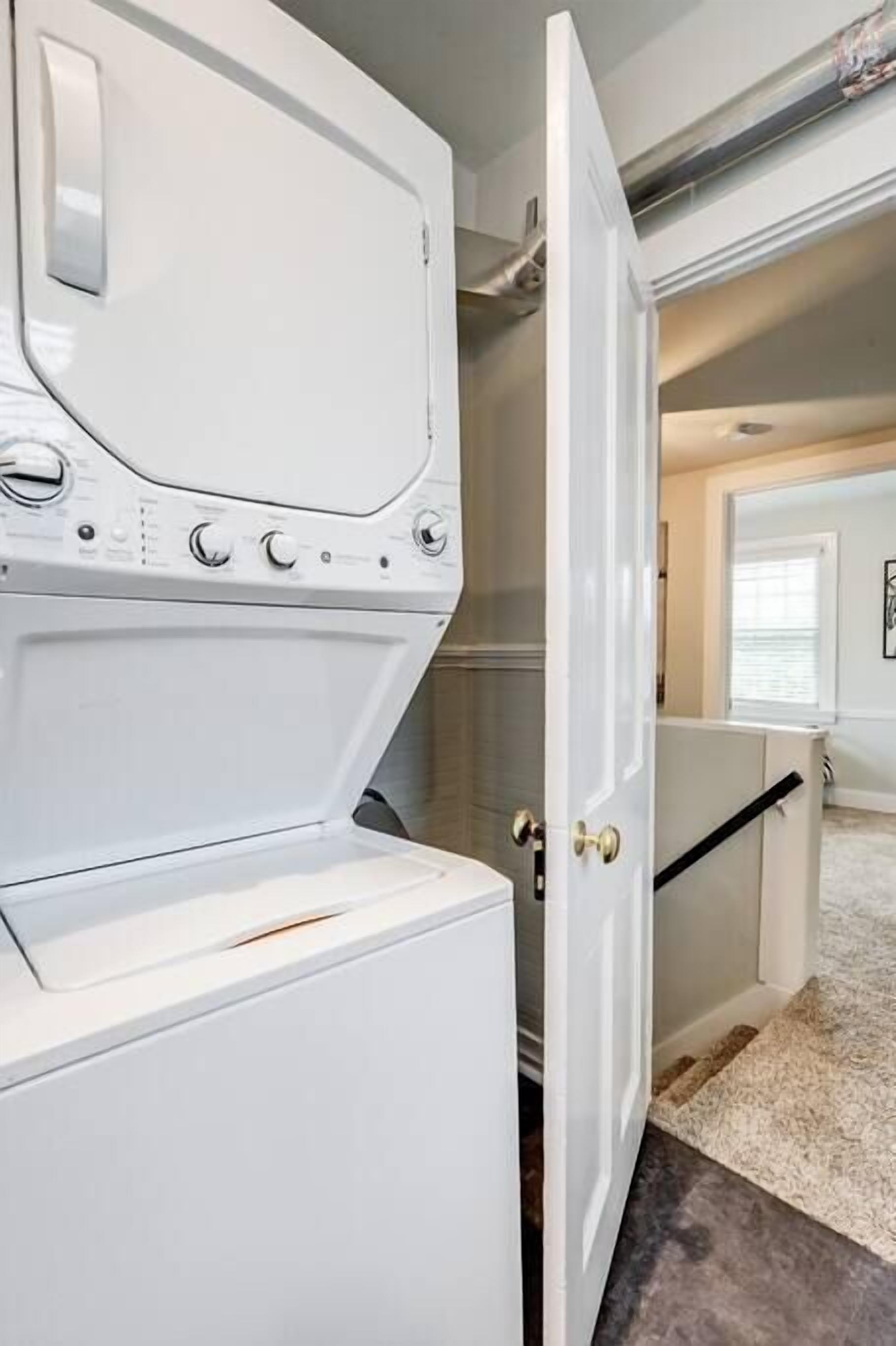 A laundry room with a white washer and dryer, a door leading to another room, and a carpeted floor.