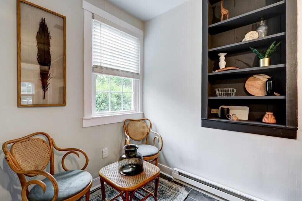 A room with two wicker chairs, a small table with a vase, a bookshelf with various items, and a framed feather on the wall.