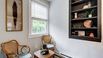 A room with two wicker chairs, a small table with a vase, a bookshelf with various items, and a framed feather on the wall.