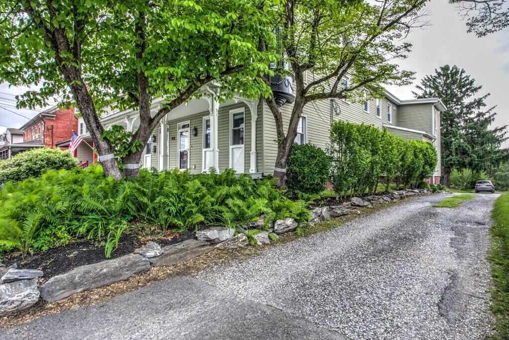 A two-story house with a front porch, surrounded by lush greenery and a gravel driveway.