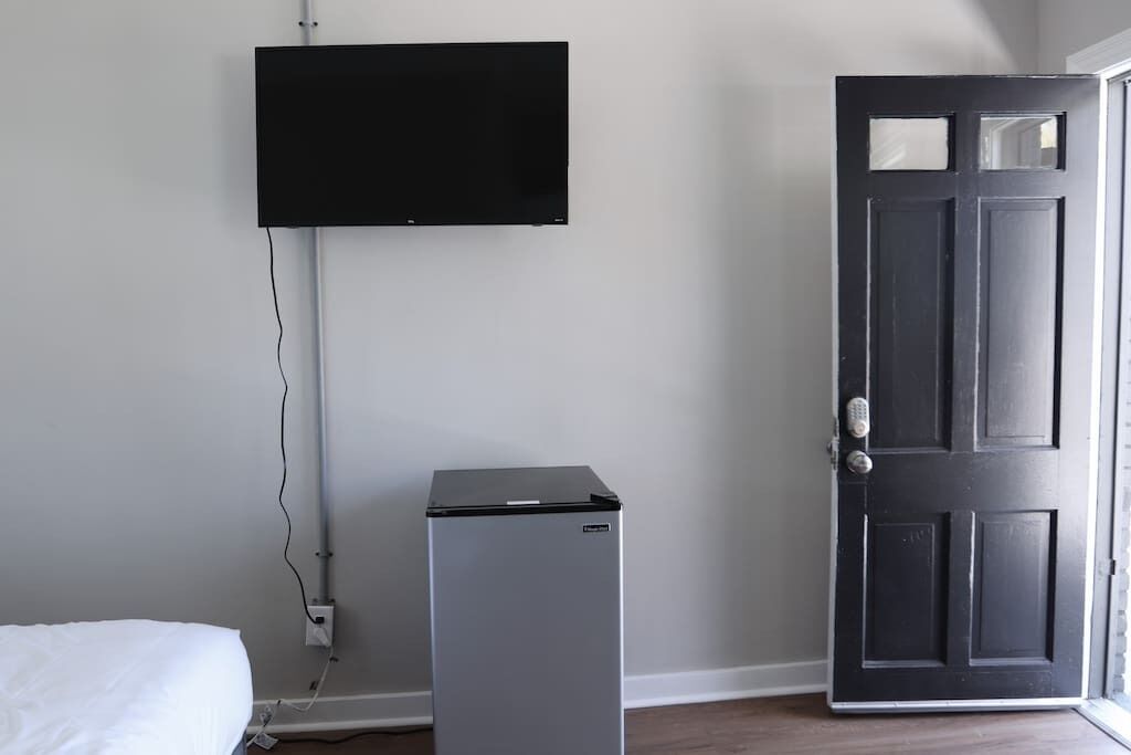 Una habitación con una cama, un refrigerador pequeño, una puerta y un televisor montado.