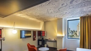 A hotel room with a modern design, featuring a yellow sofa, red chairs, a round table, a flat-screen TV, and a city view through a large window. - Ibis Baku City Hotel (Baku)