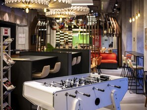 Una mesa de fútbolín en un ambiente interior moderno con zonas de estar y un letrero visible de 'ibis', indicando un hotel o establecimiento similar.