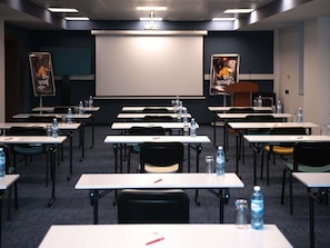 Une salle de conférence avec des rangées de tables, de chaises et un écran de projection.