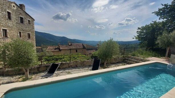 A swimming pool with a slide, surrounded by stone buildings and a mountainous backdrop. - Characteristic stone house (Le Poët-Laval)