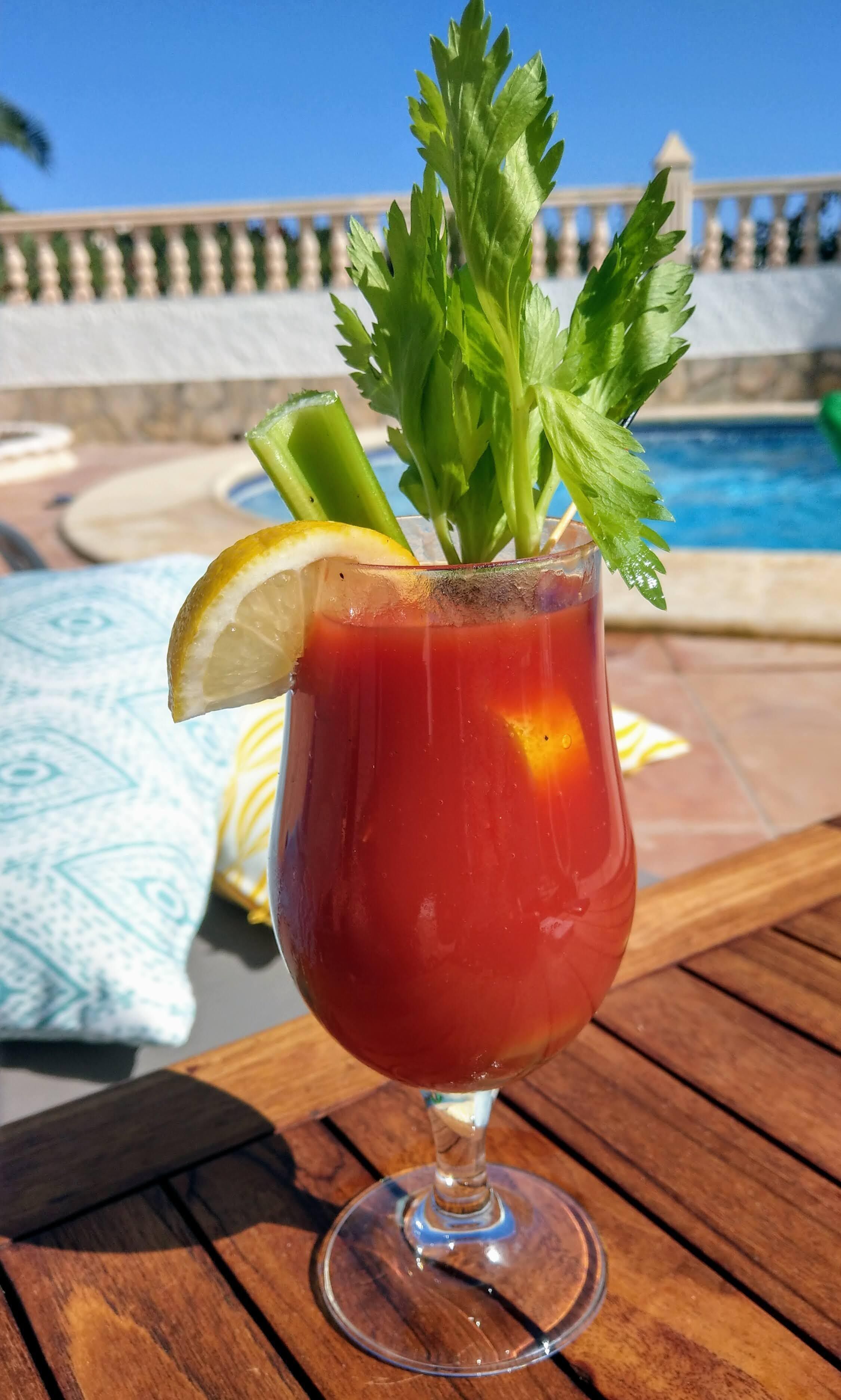 A Bloody Mary cocktail with celery and lemon garnish, placed on a wooden table near a pool.