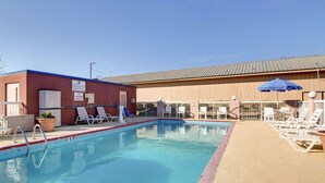 A swimming pool with lounge chairs and umbrellas, a lifeguard sign, and a building with a metal roof. - Quality Inn Brookhaven, MS (Brookhaven)