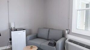 Een knus woongedeelte met een grijze bank, een klein rond tafeltje met een plant, een witte minibar en een raam met uitzicht.