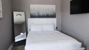 Een net opgemaakt bed met witte lakens, een grote spiegel aan de linkerkant, en een schilderij boven het bed.