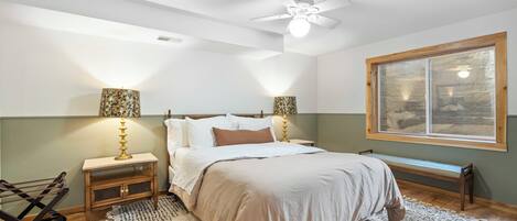Un dormitorio con cama, mesitas de noche, ventilador de techo, ventana y un banco.
