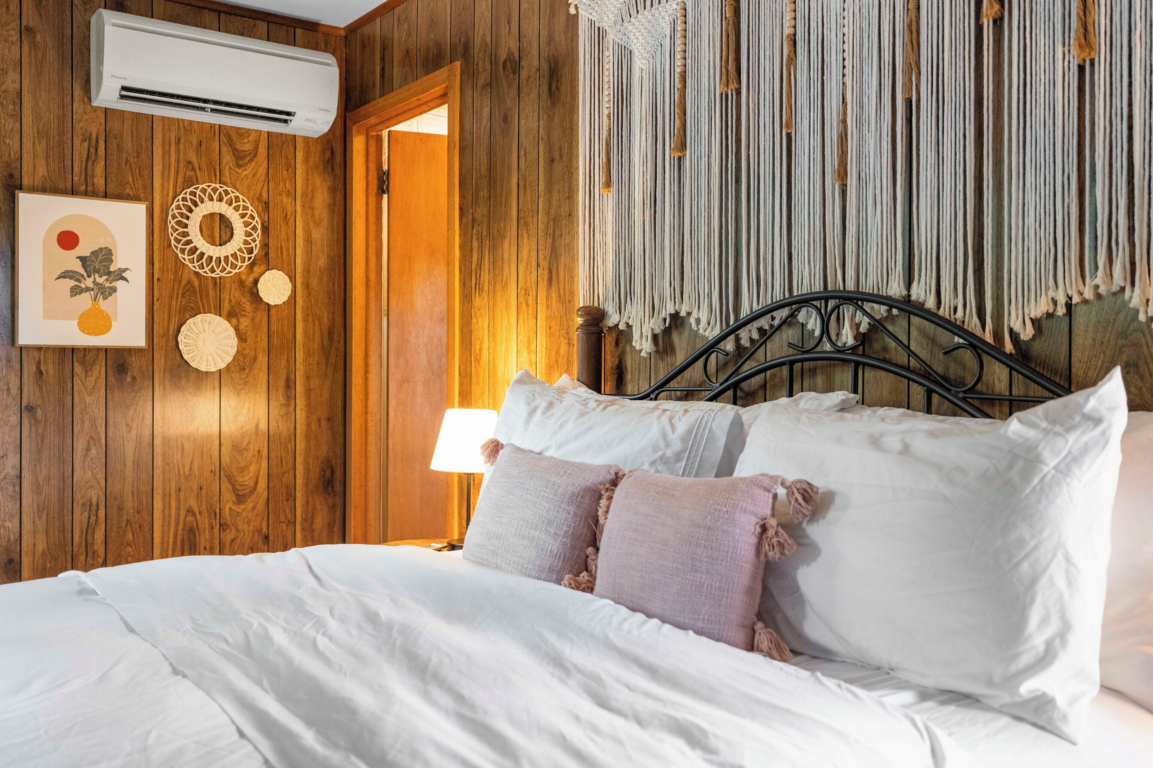 A wooden room with a bed, an air conditioner, a macrame wall hanging, and a door.